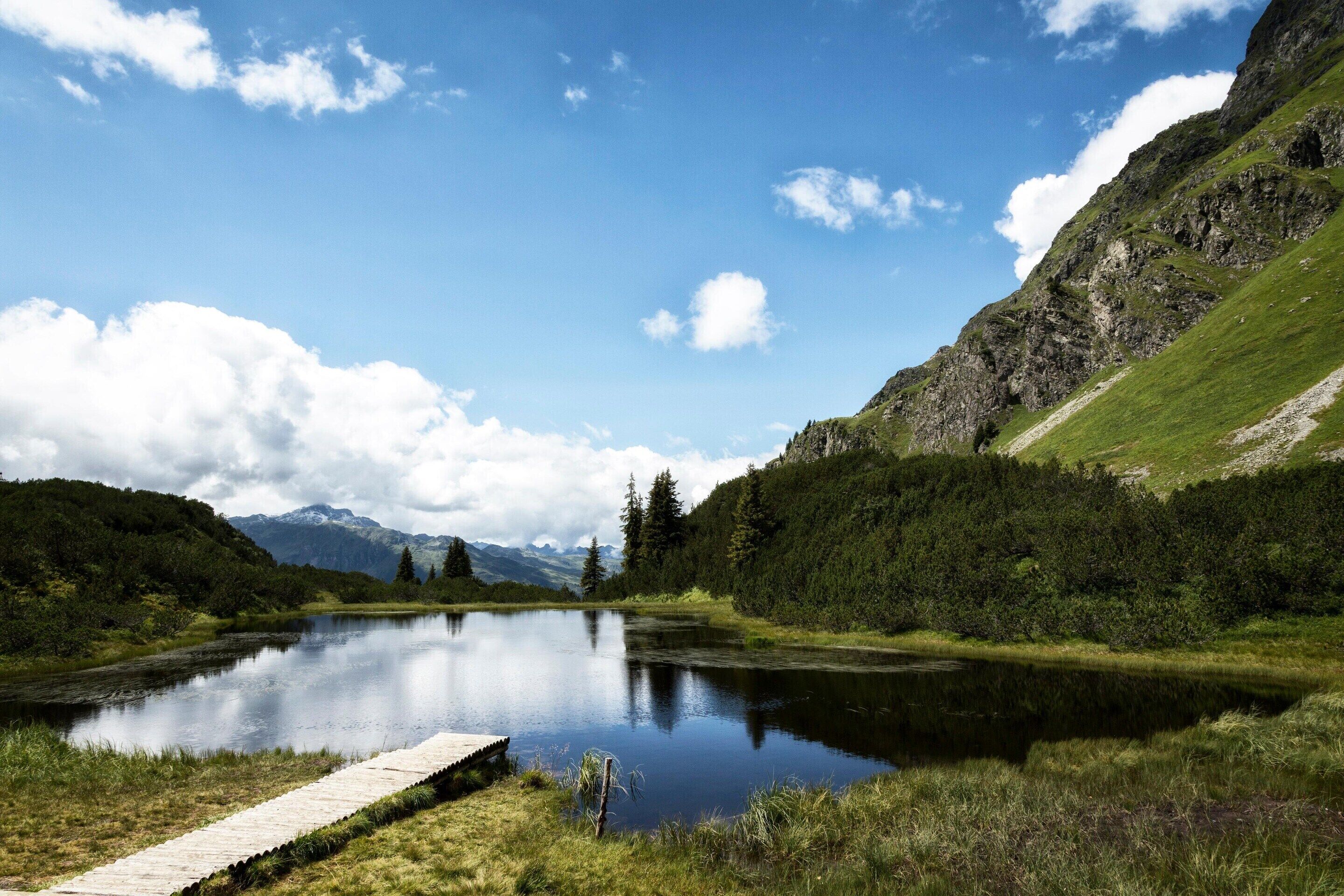 A tranquil lake with a wooden dock, surrounded by lush greenery and mountains in the distance.