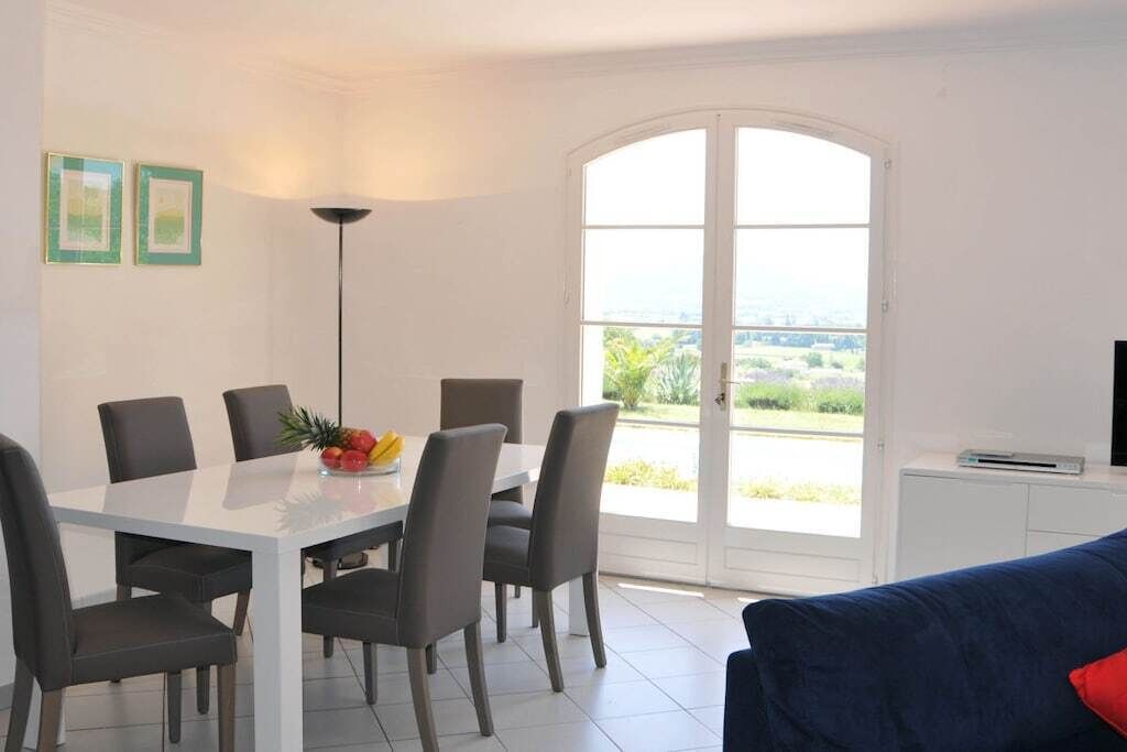 Un coin repas avec une table et des chaises blanches, un panier de fruits et une vue à travers une porte vitrée.