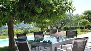 An outdoor dining area with a glass table and chairs, surrounded by lush greenery and a view of a swimming pool.