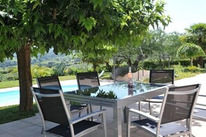 Un espace de restauration en plein air avec une table et des chaises en verre, entouré d’une végétation luxuriante et offrant une vue sur une piscine.