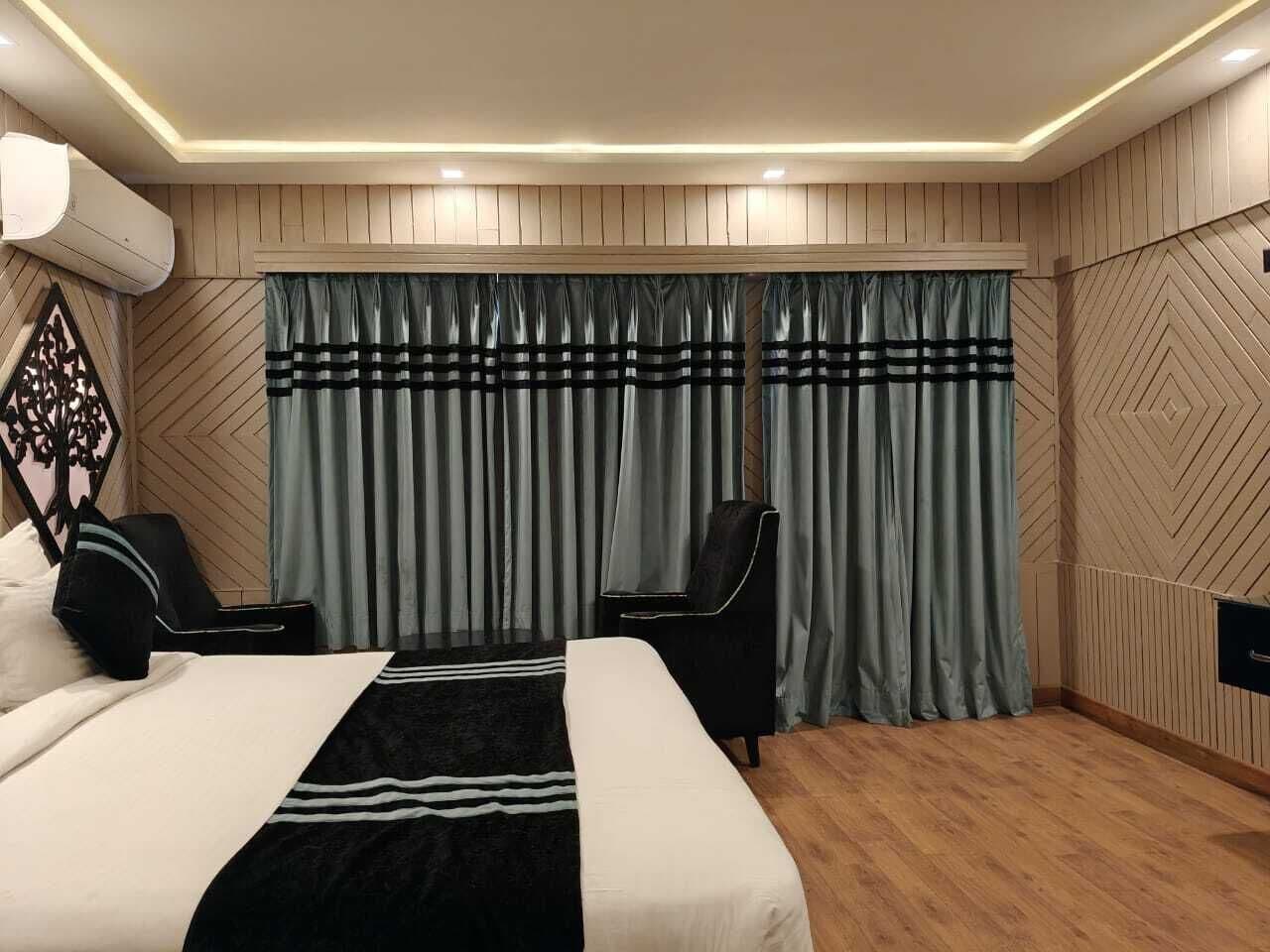A hotel room with a bed, two chairs, a large window with curtains, and a wall-mounted air conditioning unit.