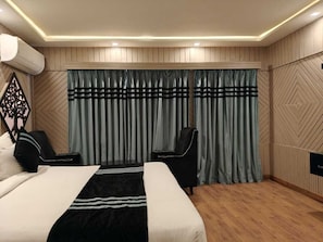 A hotel room with a bed, two chairs, a large window with curtains, and a wall-mounted air conditioning unit. - The Heritage Kud (Patnitop)