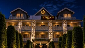 Un hotel de varios pisos con un letrero llamativo que dice 'The Heritage Kud'. Cuenta con balcones con elaborados diseños de celosía y una fachada simétrica.