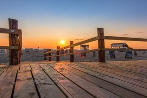 Ein hölzerner Steg führt zu einem Strand, im Hintergrund ist ein Sonnenuntergang zu sehen.