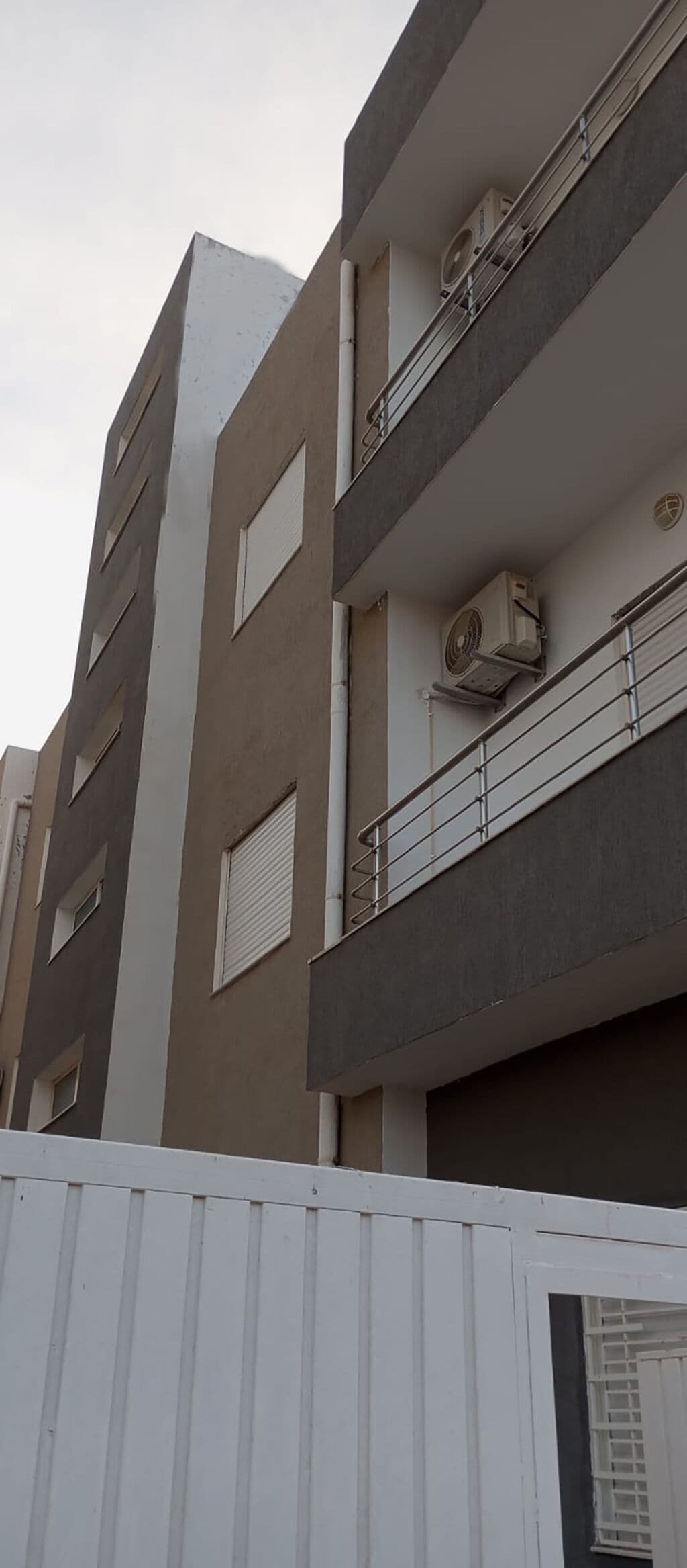 A multi-story building with balconies, air conditioning units, and a white fence in the foreground.