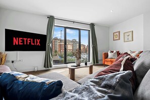 Ein modernes Wohnzimmer mit großem Fenster, einem wandmontierten Flachbildfernseher und einer gemütlichen Sitzecke mit Sofa und Sesseln.