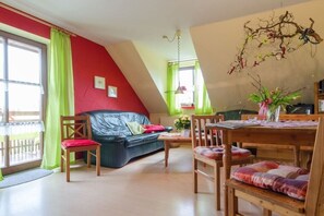 A living room with a black leather sofa, wooden dining table, and chairs. There is a window with green curtains, a hanging plant, and a wall-mounted branch decoration. - Cherry Orchard at Haus Bayer (Pottenstein)