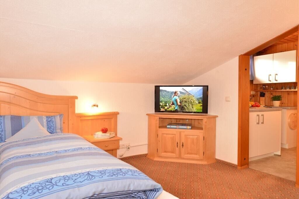 A bedroom with a bed, a television, a wooden cabinet, and a kitchenette area.