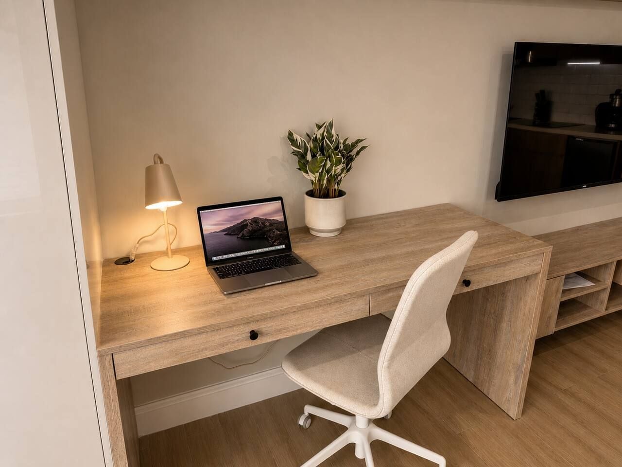 Ein moderner Arbeitsplatz mit einem Holztisch, einem Laptop, einer Schreibtischlampe, einem Topf mit einer Pflanze und einem an der Wand montierten Fernseher.