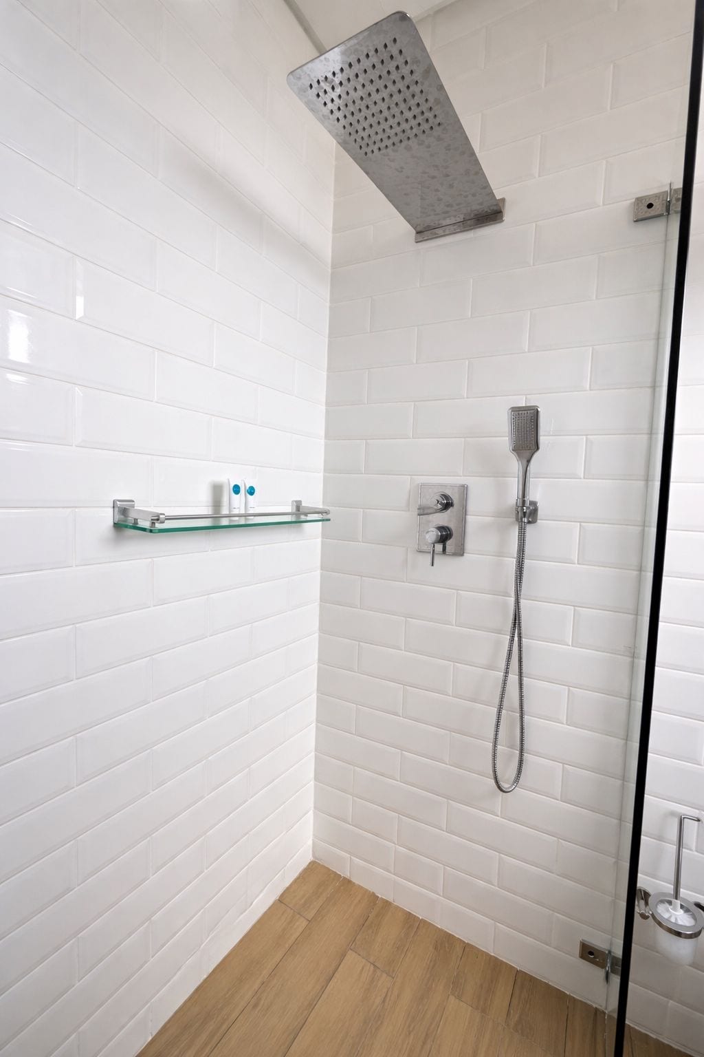 Zona de ducha con puerta de cristal, paredes con azulejos blancos y suelo de madera.