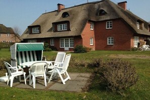 Een rieten dak huis met een bakstenen gevel, een terras met witte kunststof meubels en een groen zonnescherm.