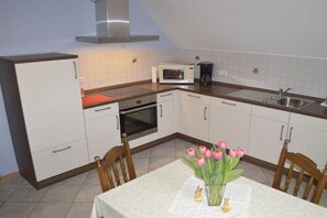 Une cuisine compacte avec des meubles blancs, un four à micro-ondes, une cuisinière et un évier. On y trouve une table à manger avec des chaises et un vase de fleurs.
