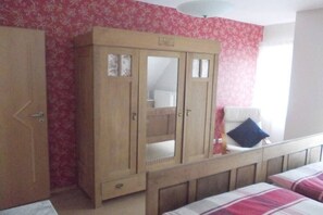 Une chambre avec une armoire en bois, un lit recouvert d’une couverture à carreaux et une fenêtre avec un rideau blanc.