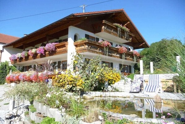 Un edificio de dos pisos con balcones decorados con flores, un estanque pequeño y mesas al aire libre.