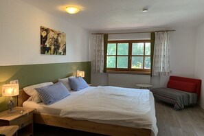 Un dormitorio con cama, lámparas de noche, una ventana con cortinas, un banco rojo y un cuadro en la pared.