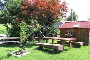Una mesa de picnic con bancos, una pequeña estructura de madera con techo rojo y un árbol de hojas rojas.