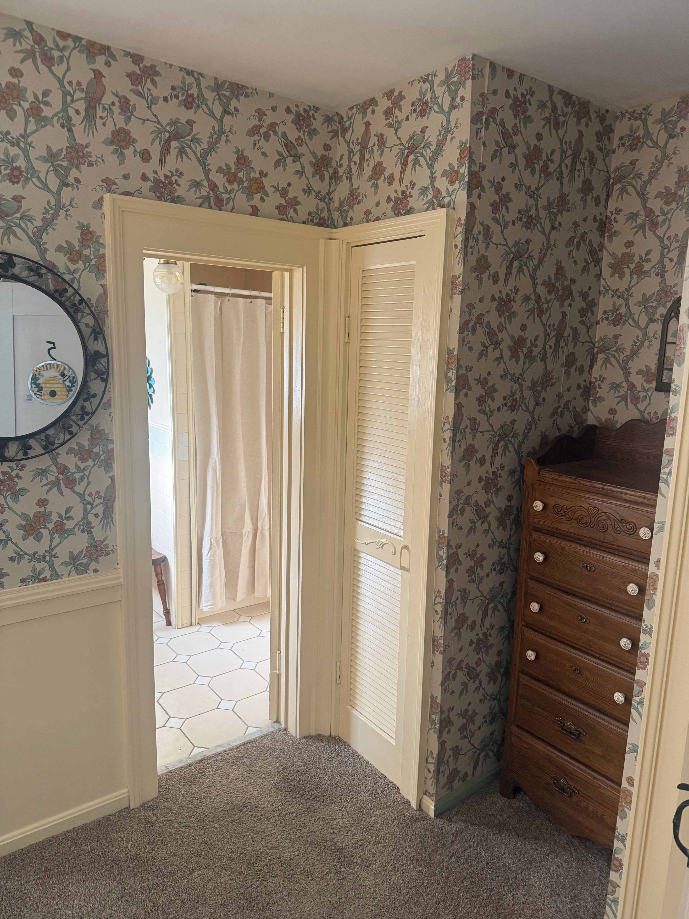 A room with floral wallpaper, a wooden dresser, and a door leading to a bathroom with a shower curtain.