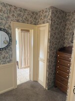 Una habitación con papel tapiz floral, un tocador de madera y una puerta que da a un baño con cortina de ducha.
