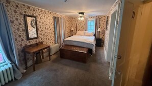 A bedroom with a bed, a wooden bench, a radiator, and a window with curtains. - Historic Riders Hostelry (Painesville)