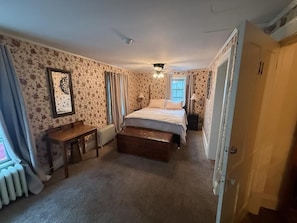 A bedroom with a bed, a wooden bench, a radiator, and a window with curtains. - Historic Riders Hostelry (Painesville)
