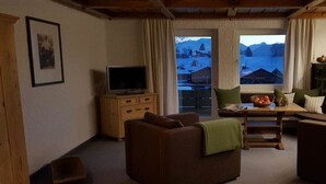 Uma sala de estar aconchegante com sofá, televisão, um armário de madeira e janela com vista para uma paisagem nevada.