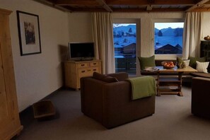 A cozy living room with a sofa, a television, a wooden cabinet, and a window offering a view of a snowy landscape. - Holiday apartment with 2 bedrooms (Sonthofen)
