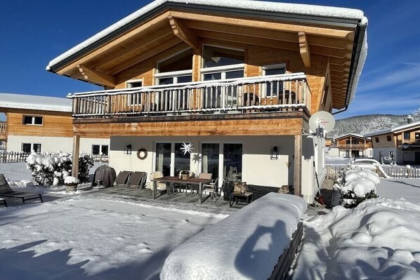 A wooden chalet with a balcony, outdoor seating, and snow-covered surroundings.