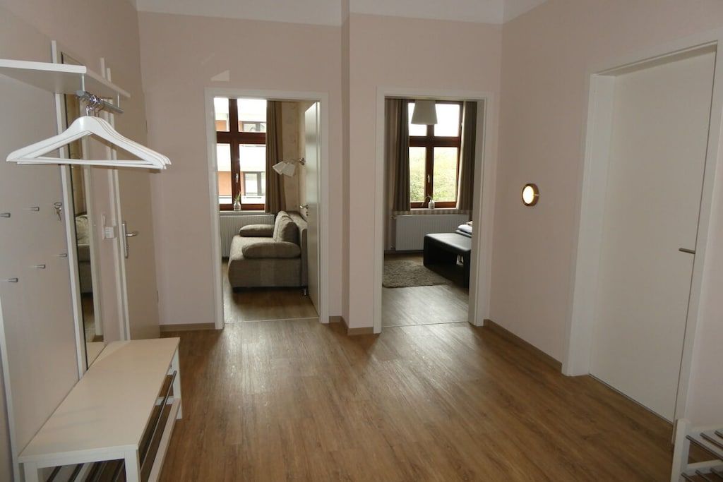 A room with a wooden floor, a white bench, a hanging coat rack, and a door leading to another room.