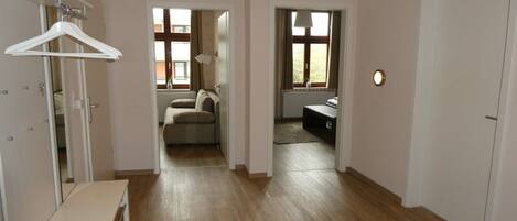 A room with a wooden floor, a white bench, a hanging coat rack, and a door leading to another room.