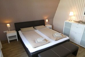 A neatly made bed with pillows and folded towels, a bedside table with a lamp, and a bench at the foot of the bed. - Holiday apartment with 4 bedrooms (Wangerooge)