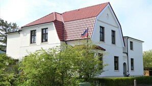 A two-story white building with a red-tiled roof, a flag, and a green utility box. - Holiday apartment with 4 bedrooms (Wangerooge)