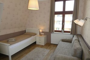 Een slaapkamer met een enkel bed, een slaapbank, een nachtkastje met een lamp, een radiator en een raam met gordijnen.