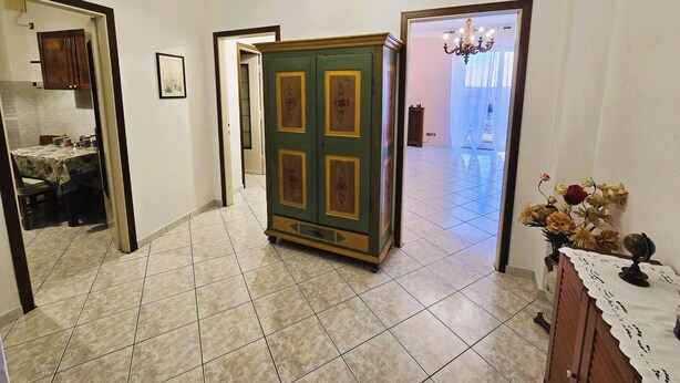 A hallway with a tiled floor, a green cabinet with decorative panels, a dining area with a table and chairs, and a chandelier in the adjacent room. - Galileo Ferraris Apartment, Short-Term Rentals in the City of Vercelli (Vercelli)