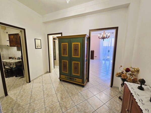 Un corridoio con pavimento piastrellato, un armadietto verde con pannelli decorativi, una sala da pranzo con tavolo e sedie, e un lampadario nella stanza adiacente.