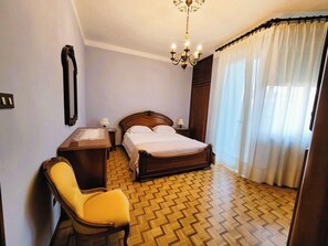 A hotel room with a wooden bed, a desk with a chair, a chandelier, and a large window with curtains. - Galileo Ferraris Apartment, Short-Term Rentals in the City of Vercelli (Vercelli)