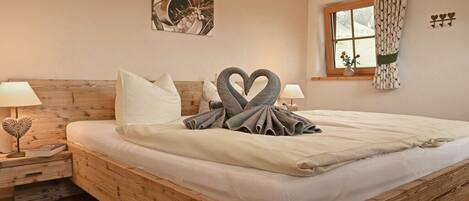 A wooden bed with a heart-shaped pillow arrangement, a bedside table with a lamp, a window with curtains, and a wall-mounted picture.