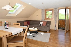 A cozy living room with a sofa, a dining table, and a small coffee table. There is a window with curtains, a skylight, and a view of greenery outside. - Wiesenblick - Family Herb (Rettenberg)