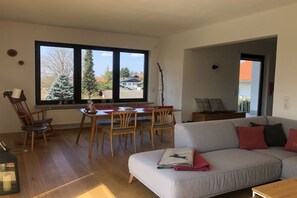A living room with a sofa, a dining table with chairs, and a large window with a view of trees and buildings. - Holiday apartment with 2 bedrooms (Chieming)