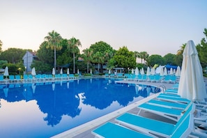 Piscina cristalina con sillas de descanso azules y sombrillas, rodeada de vegetación y edificios.