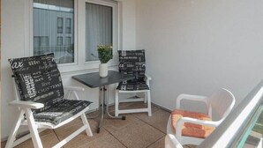 En balkong med två vita stolar och ett bord, en krukväxt och ett fönster med gardiner.