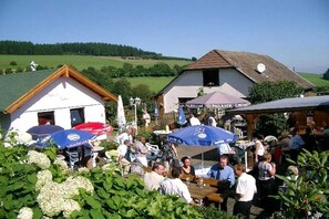 Ein Außenbereich mit Tischen und Sonnenschirmen, umgeben von Grünflächen und Gebäuden.