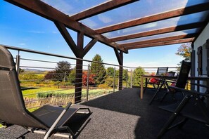 Een overdekt terras met een tafel en stoelen, met uitzicht op een tuin, en een gebouw op de achtergrond.