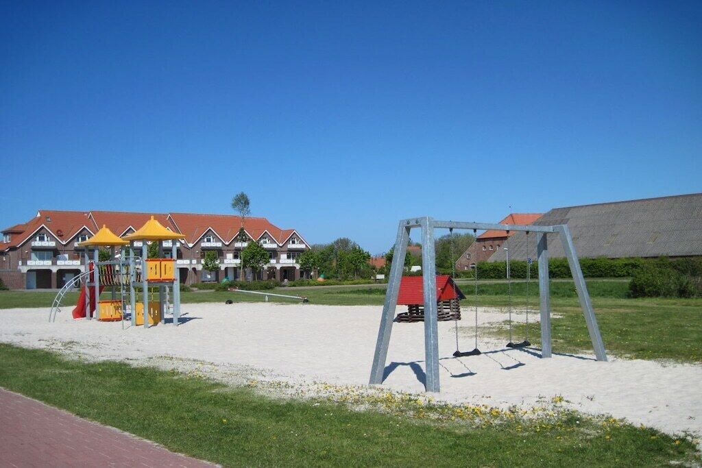 A playground with a swing set and climbing structures, a sandy area, and a building with a red roof in the background.