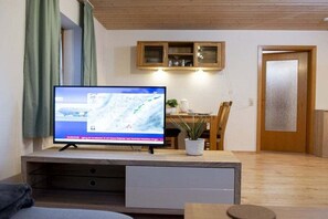 Een televisie op een houten standaard in een kamer met een houten plafond, een eettafel met stoelen en een kast boven de tafel.