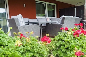 Un espace extérieur aménagé avec des meubles en osier et une table, entouré de verdure et de fleurs rouges.