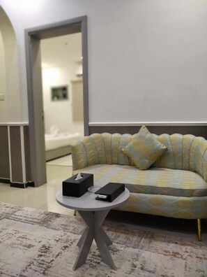 A room with a sofa, a small round table with a tissue box and remote control, and a patterned rug. - Alhalm Aljamel Serviced Apartments (Jeddah, Al-Samir District)