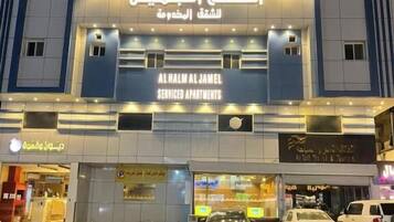 A building with a sign for 'AL HAML AL JAMEEL SERVICED APARTMENTS' and a parking area with cars.
