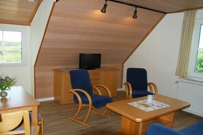 Quarto com teto inclinado, móveis de madeira, televisão sobre um móvel e janela com vista para o verde.