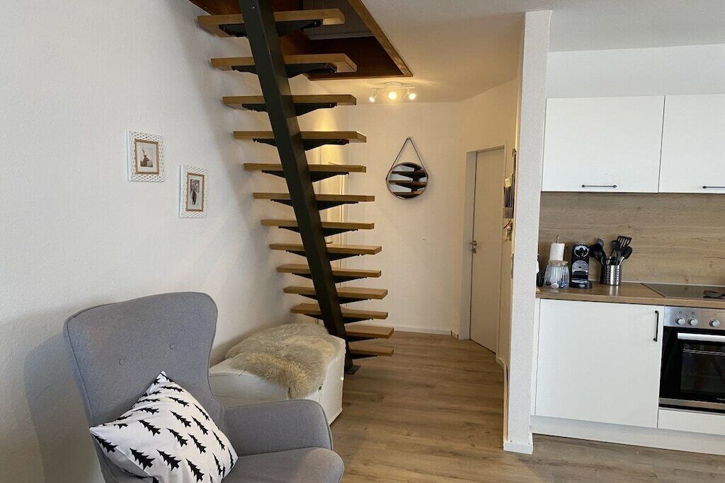 A modern kitchen with white cabinets and a wooden countertop, a grey sofa with a patterned pillow, and a spiral staircase with wooden steps.