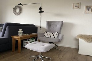 Un salon comprenant un fauteuil gris, un pouf blanc, une table d’appoint en bois, un lampadaire noir et un canapé gris.